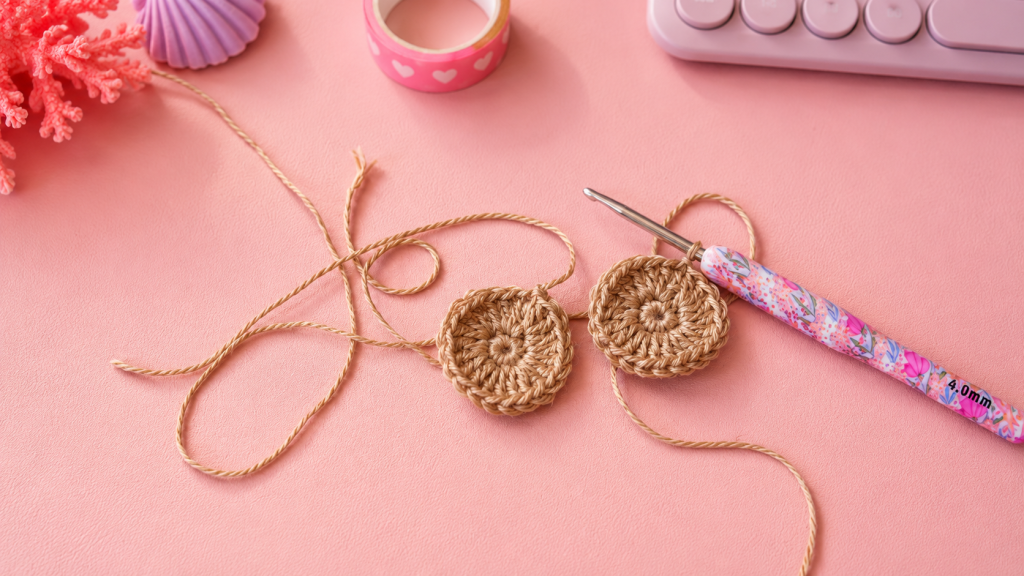 Conchiglia Nautilus uncinetto: tutorial facile per creare un tesoro del Mare Pinky 5 Due cerchi all'uncinetto realizzati con filato color sabbia e uncinetto decorato, appoggiati su uno sfondo rosa in stile Pinky Summer Time con perle, conchiglie e decorazioni marine.